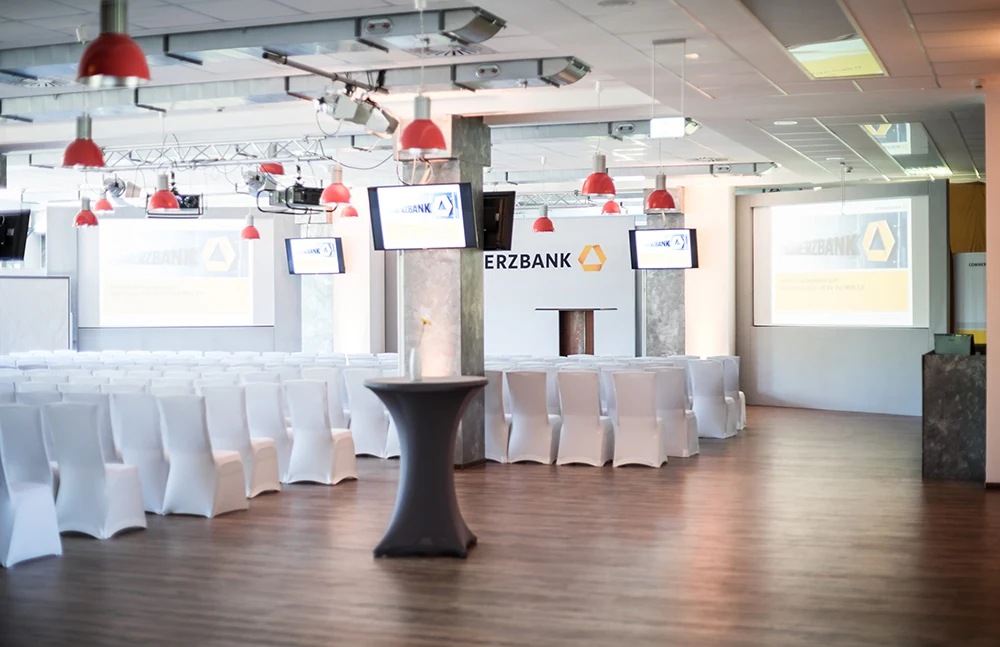Saal mit weiß eingedeckten Stehtischen und Commerzbank-Screens